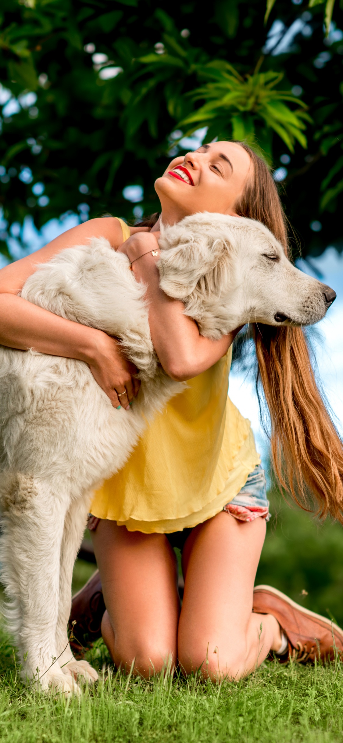 Woman in Yellow Dress Sitting on Green Grass Field With White Short Coated Dog During Daytime. Wallpaper in 1125x2436 Resolution