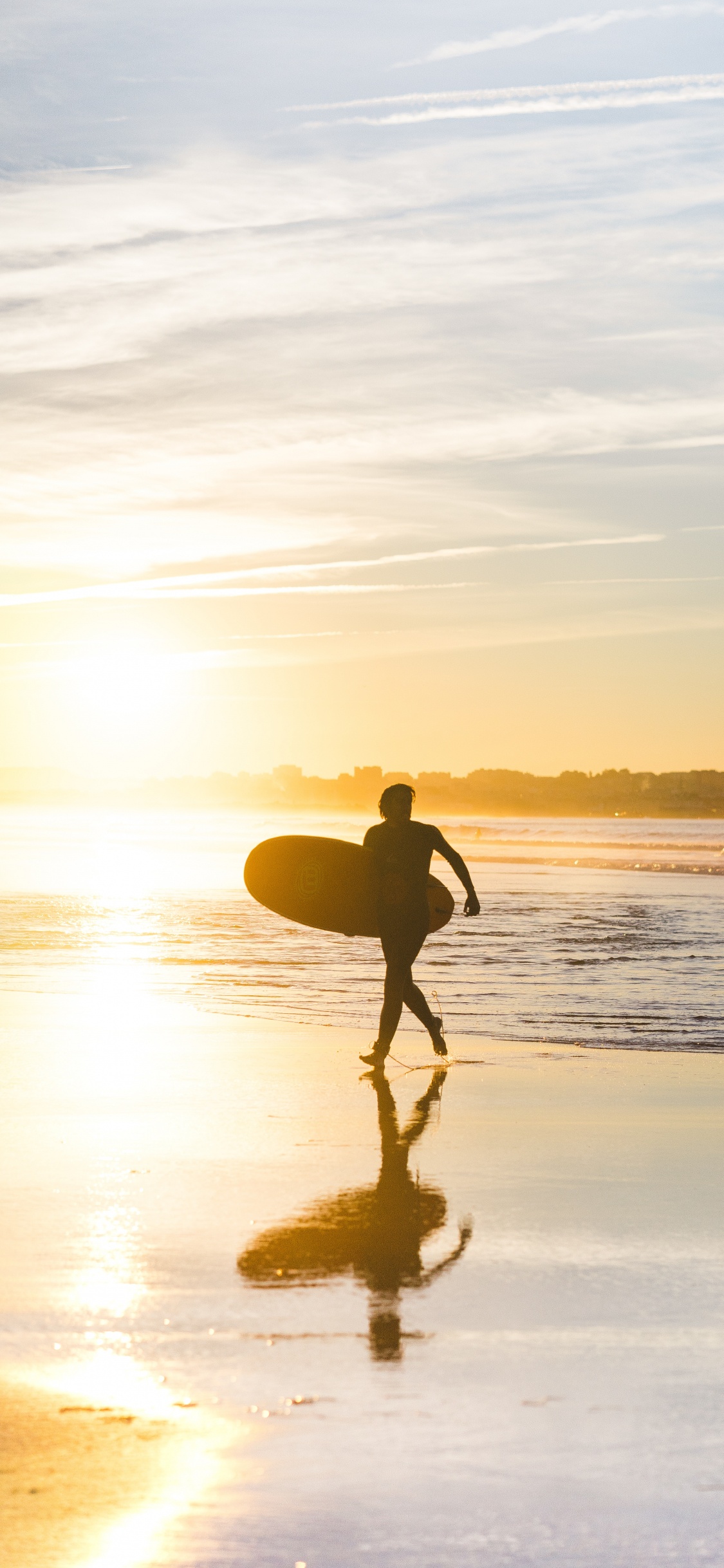 Hombre en Shorts Negros Sosteniendo la Tabla de Surf Caminando Por la Orilla Del Mar Durante la Puesta de Sol. Wallpaper in 1125x2436 Resolution