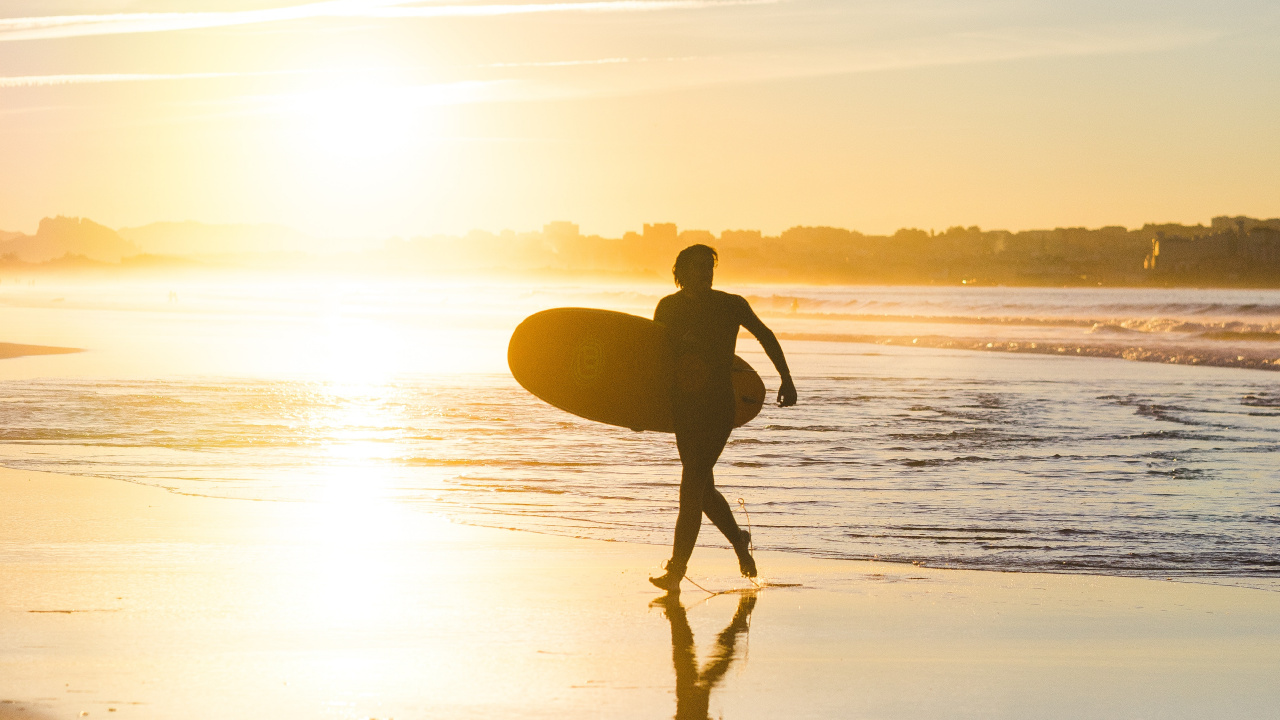 Hombre en Shorts Negros Sosteniendo la Tabla de Surf Caminando Por la Orilla Del Mar Durante la Puesta de Sol. Wallpaper in 1280x720 Resolution