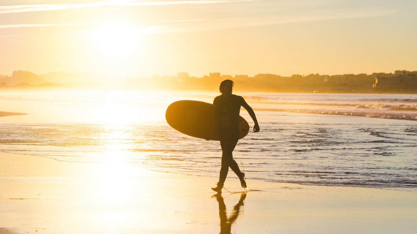 Man in Black Shorts Holding Surfboard Walking on Seashore During Sunset. Wallpaper in 1366x768 Resolution