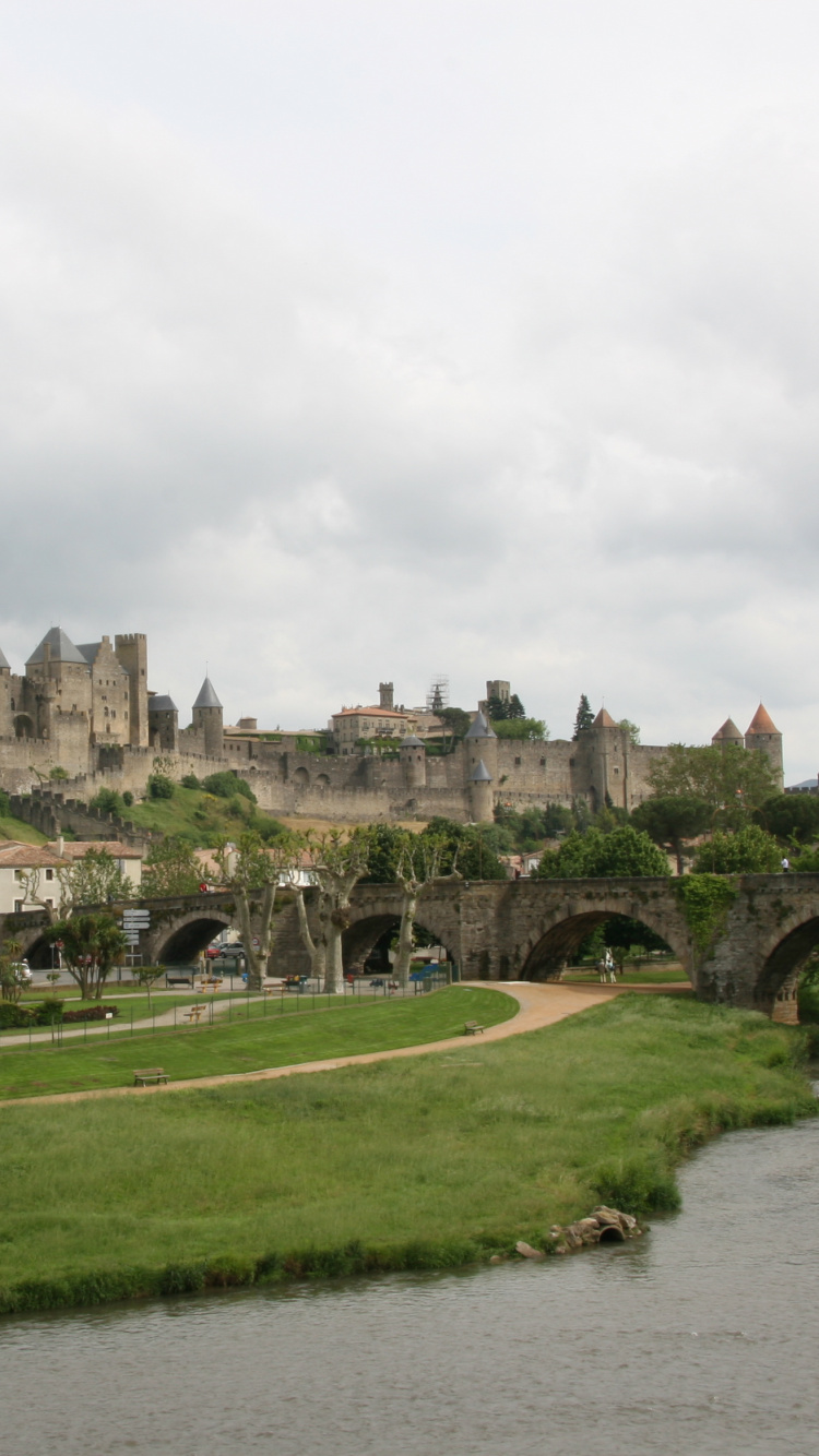 Braunes Betongebäude in Der Nähe Von Grünem Grasfeld Und Fluss Unter Weißen Wolken Tagsüber. Wallpaper in 750x1334 Resolution