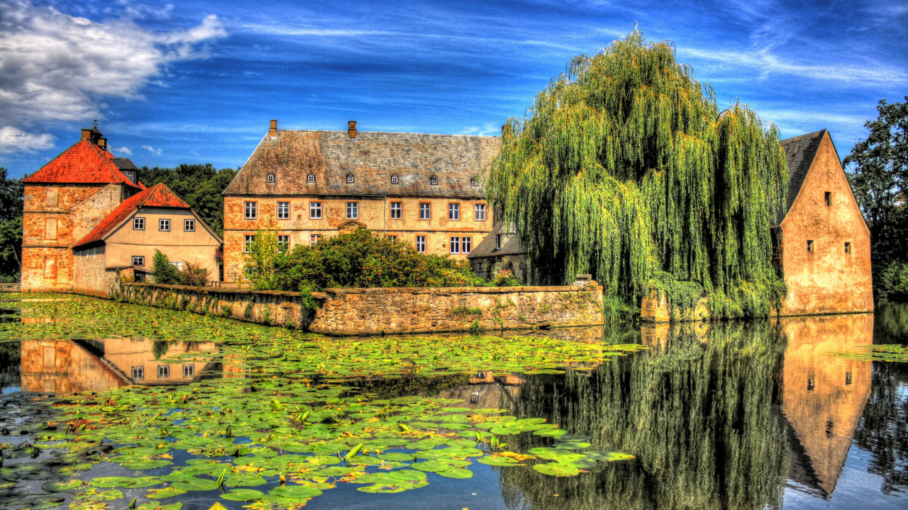 Brown Brick Building Near Green Trees and Body of Water Under Blue Sky During Daytime. Wallpaper in 1280x720 Resolution