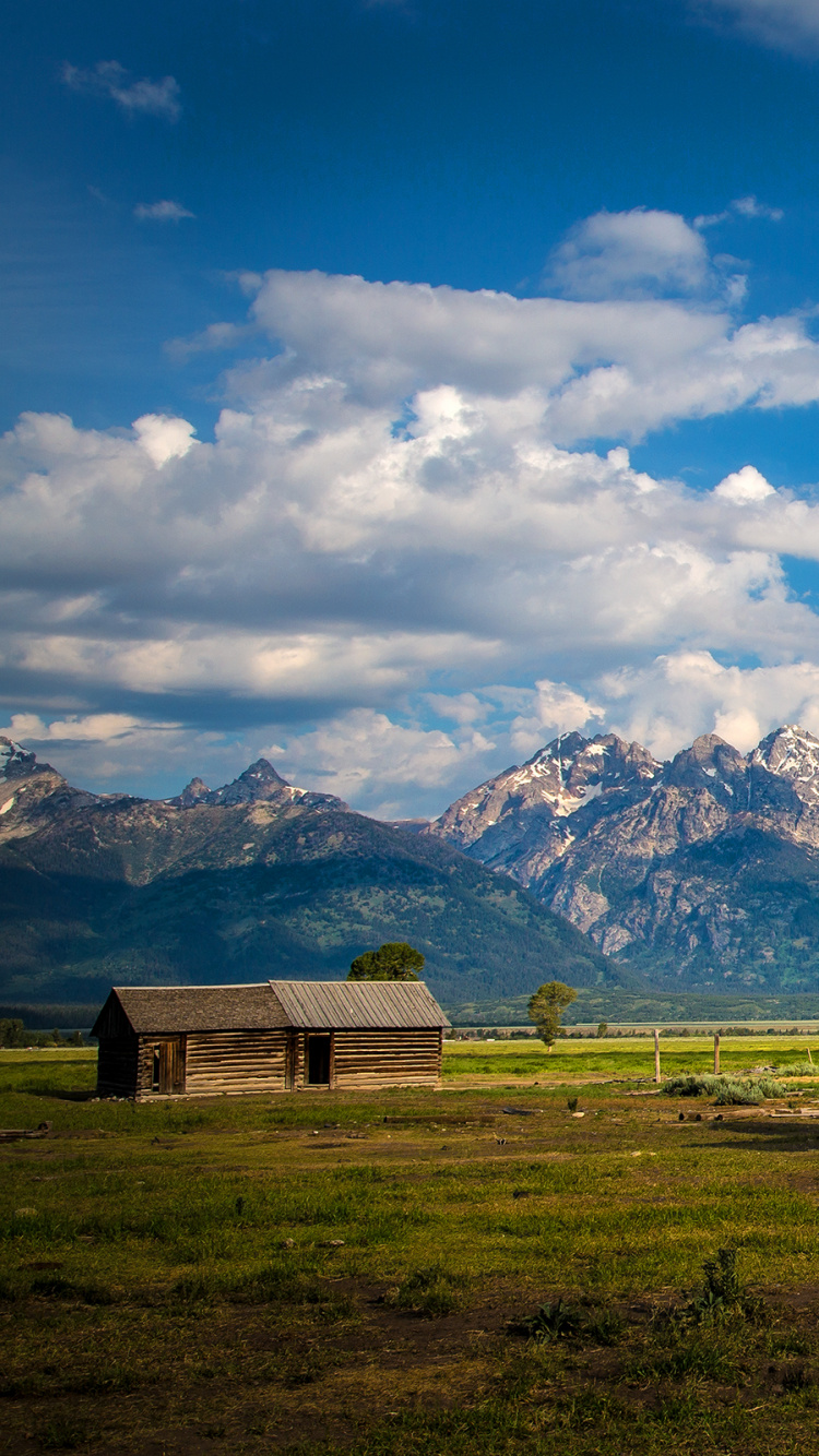 Brown Wooden Barn on Green Grass Field Near Snow Covered Mountains Under Blue and White Cloudy. Wallpaper in 750x1334 Resolution