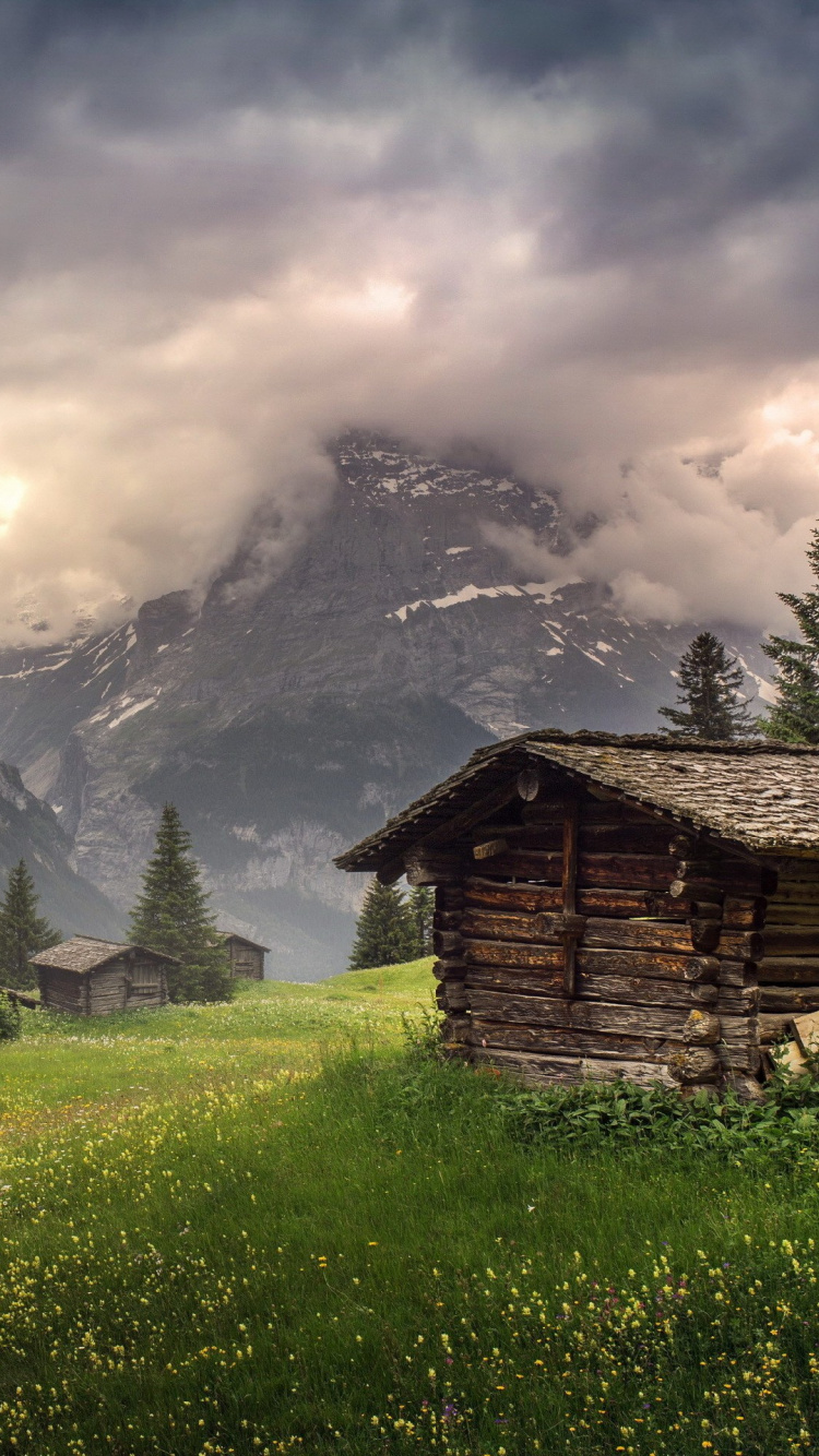 Braunes Holzhaus Auf Grünem Grasfeld in Der Nähe Des Berges Unter Weißen Wolken Tagsüber. Wallpaper in 750x1334 Resolution
