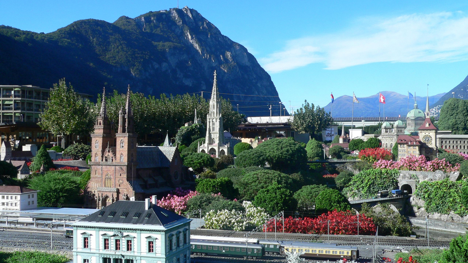 Weißes Und Braunes Betongebäude in Der Nähe Von Grünen Bäumen Und Bergen Unter Blauem Himmel Tagsüber. Wallpaper in 1920x1080 Resolution