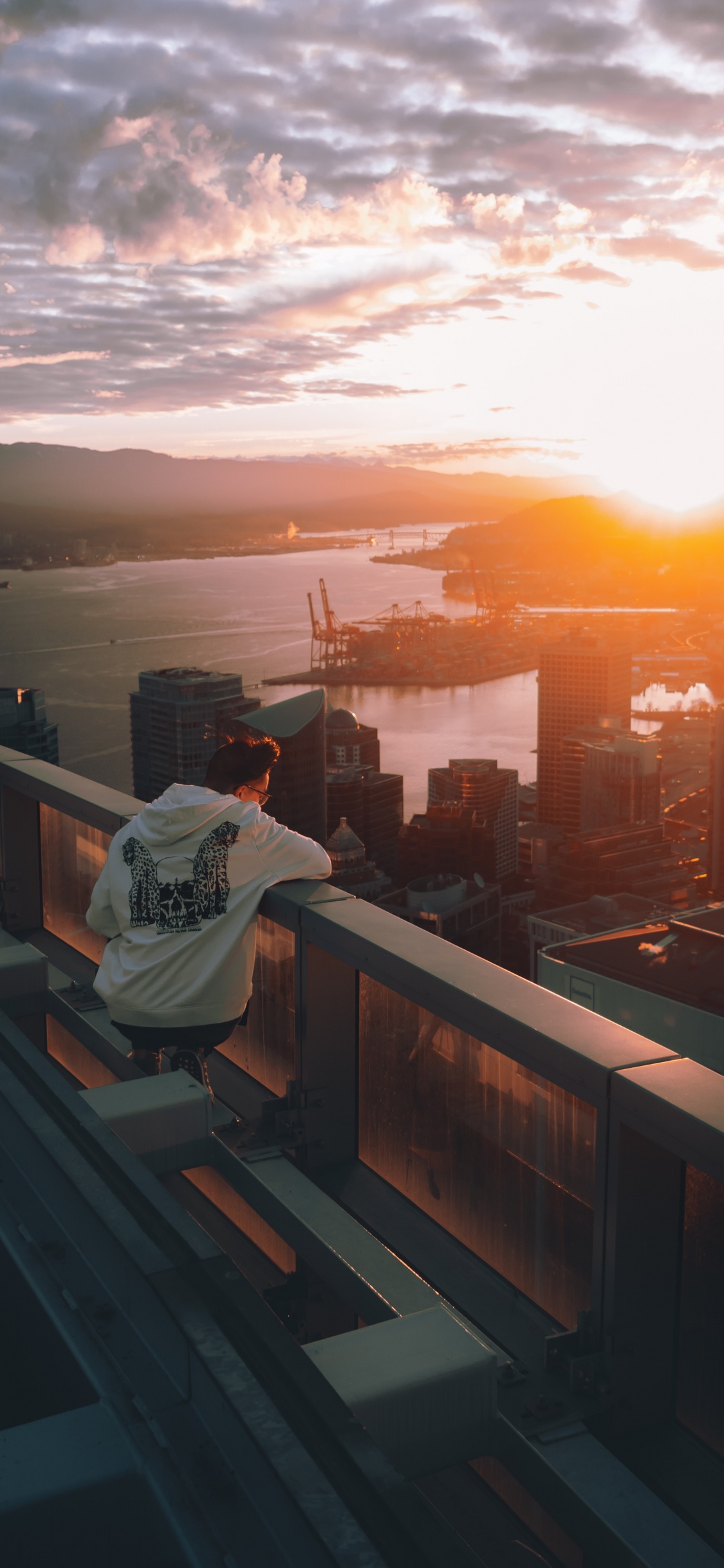Mann im Weißen Hoodie Sitzt Auf Einem Braunen Holzzaun Und Blickt Bei Sonnenuntergang Auf Die Skyline Der Stadt. Wallpaper in 1125x2436 Resolution