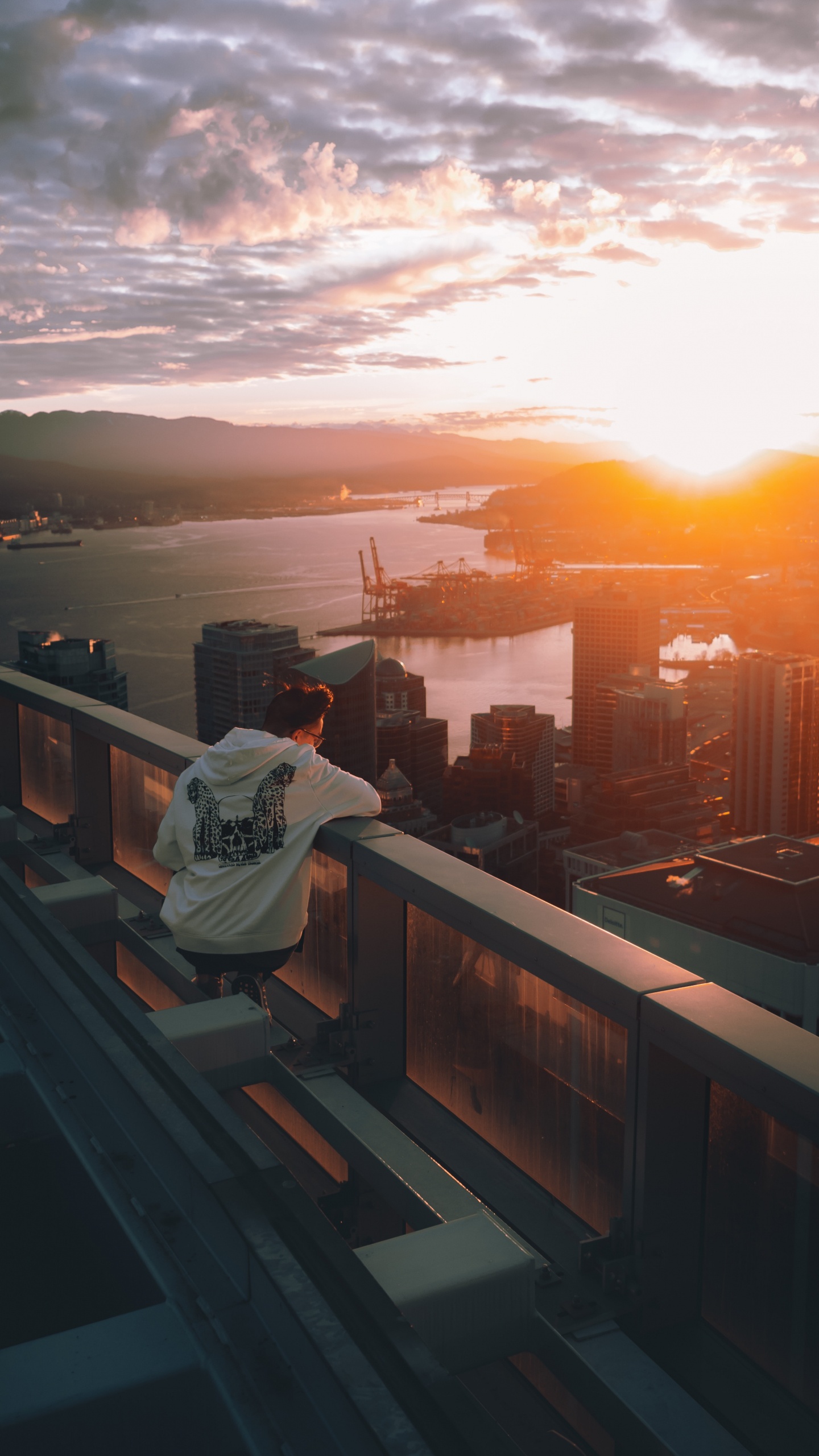 Mann im Weißen Hoodie Sitzt Auf Einem Braunen Holzzaun Und Blickt Bei Sonnenuntergang Auf Die Skyline Der Stadt. Wallpaper in 1440x2560 Resolution