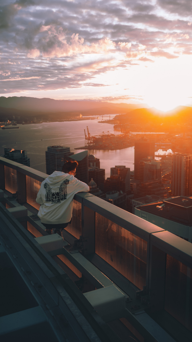 Mann im Weißen Hoodie Sitzt Auf Einem Braunen Holzzaun Und Blickt Bei Sonnenuntergang Auf Die Skyline Der Stadt. Wallpaper in 750x1334 Resolution