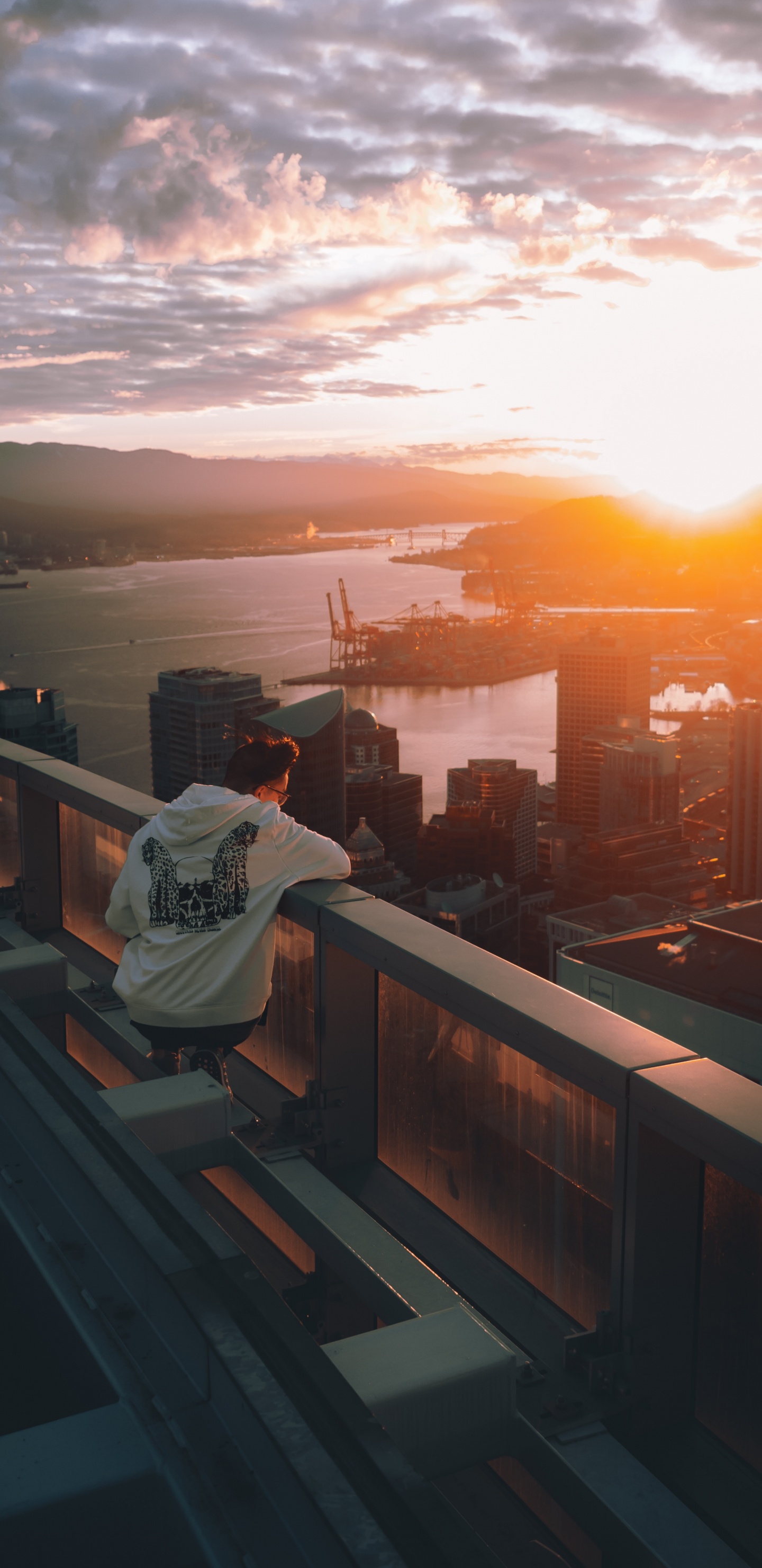 Man in White Hoodie Sitting on Brown Wooden Fence Looking at City Skyline During Sunset. Wallpaper in 1440x2960 Resolution