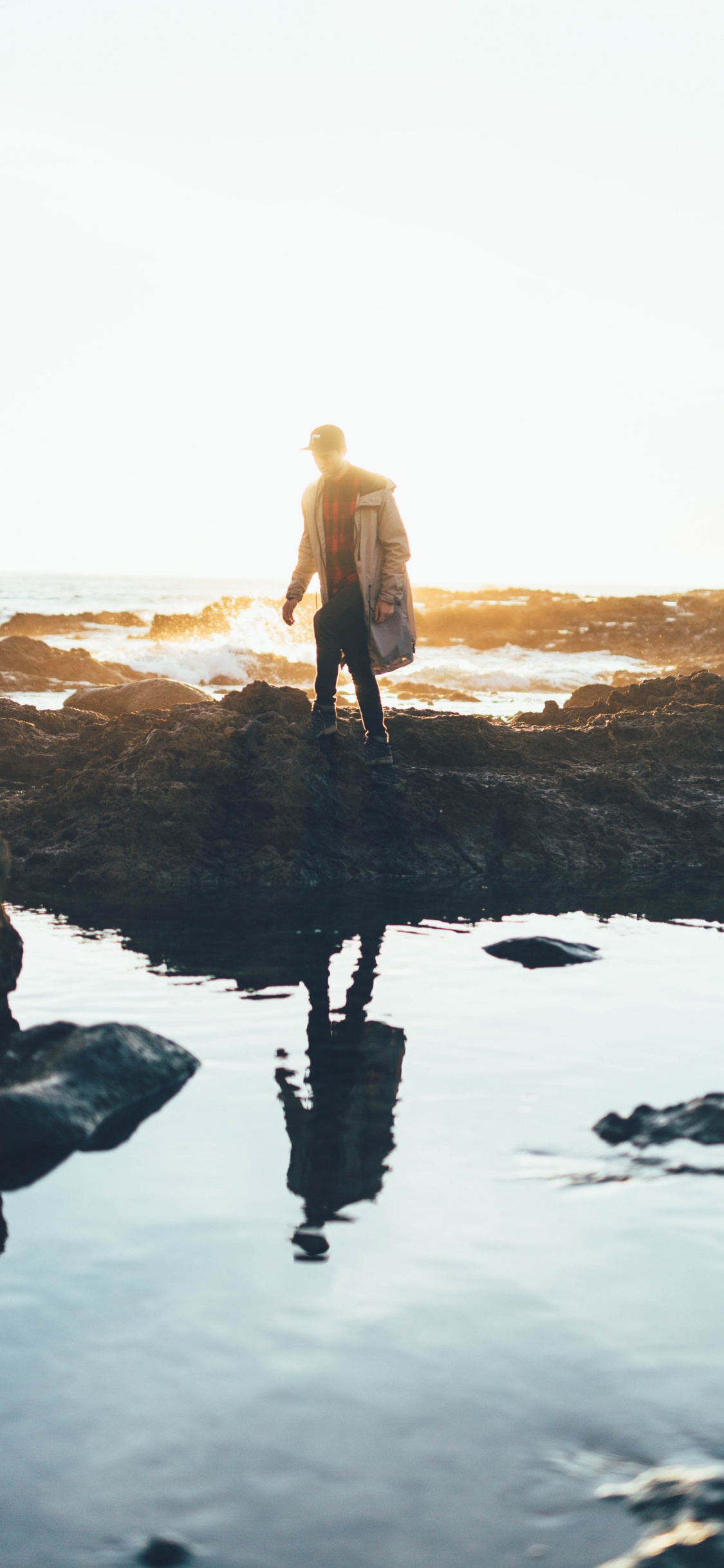 Man in Brown Coat Standing on Rock Formation Near Body of Water During Daytime. Wallpaper in 1125x2436 Resolution