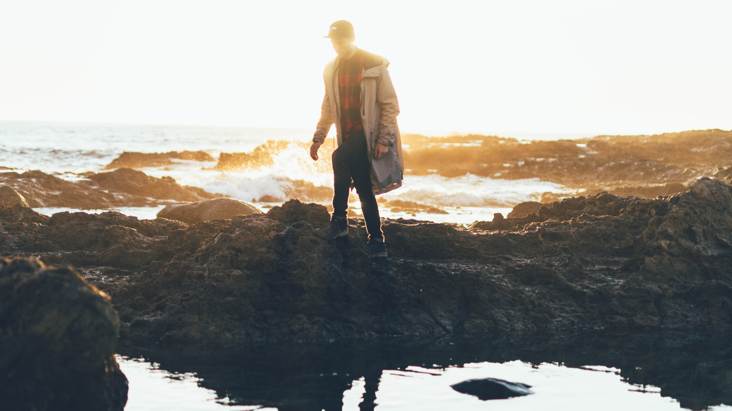 Man in Brown Coat Standing on Rock Formation Near Body of Water During Daytime. Wallpaper in 2560x1440 Resolution