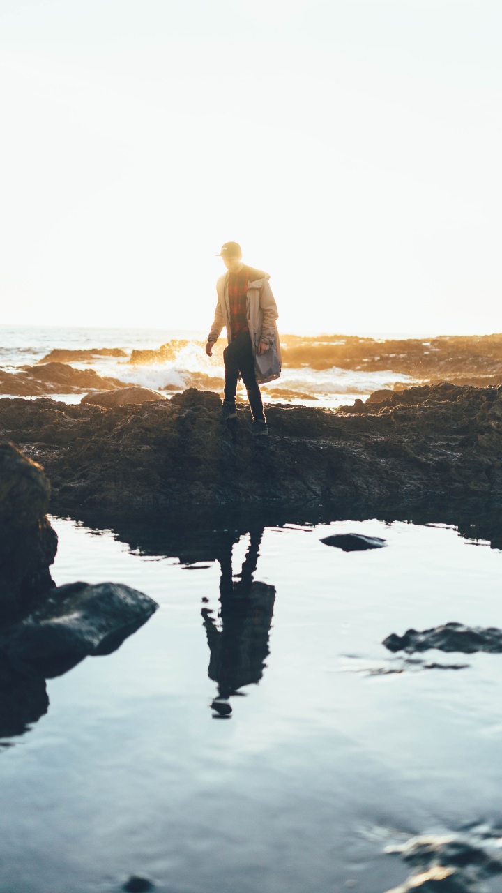 Man in Brown Coat Standing on Rock Formation Near Body of Water During Daytime. Wallpaper in 720x1280 Resolution