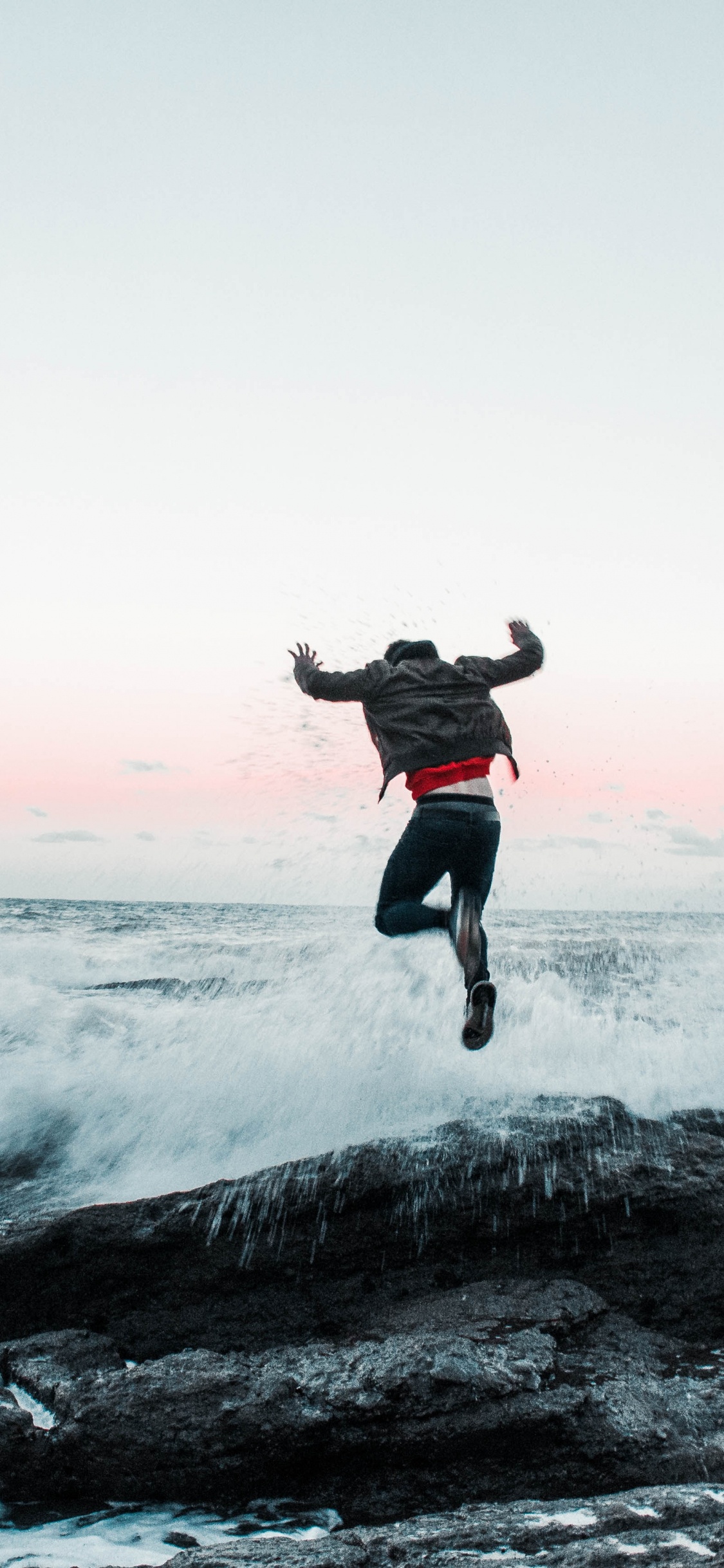 Man in Black Jacket and Pants Jumping on Rocky Shore During Daytime. Wallpaper in 1125x2436 Resolution