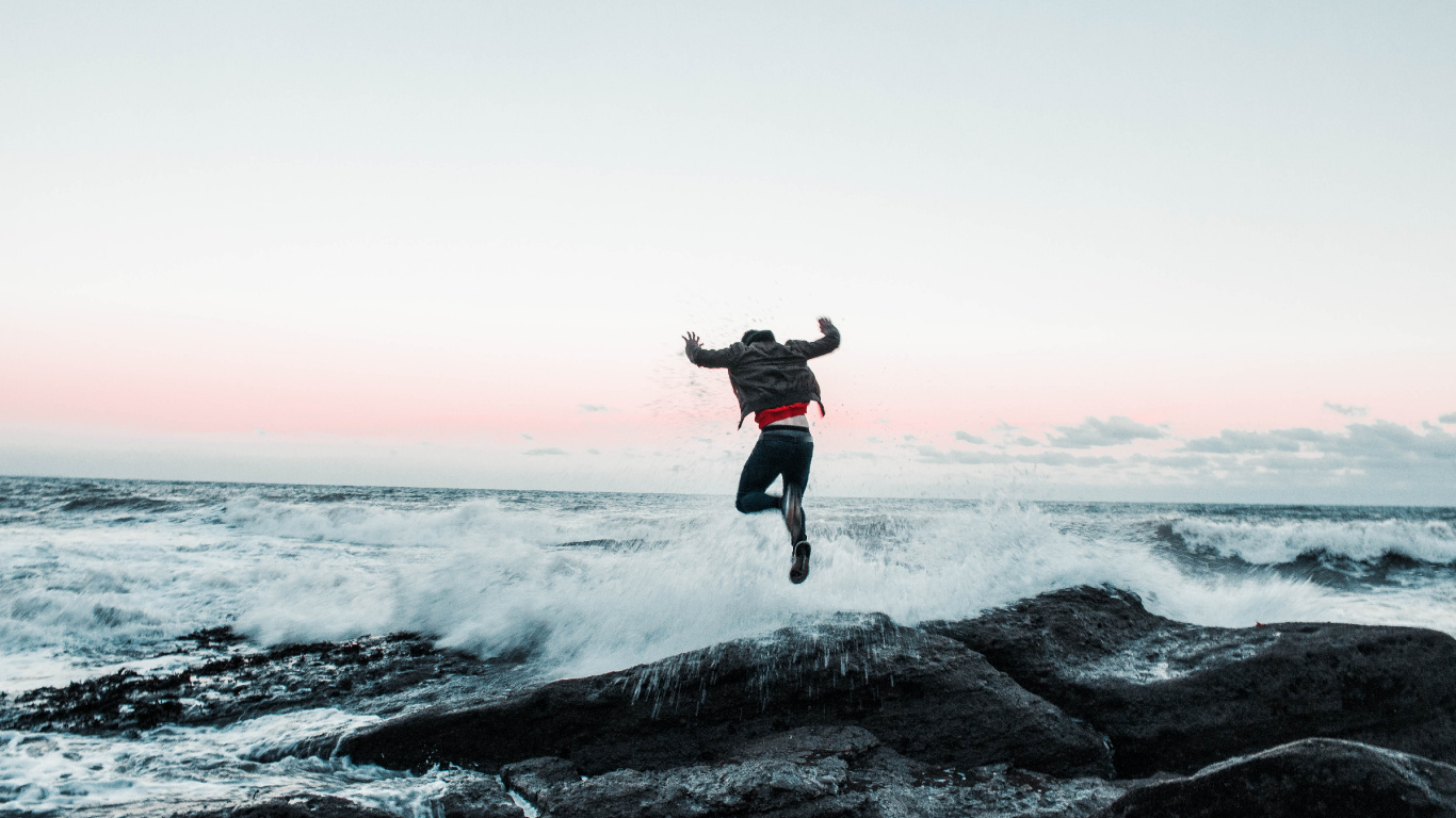 Man in Black Jacket and Pants Jumping on Rocky Shore During Daytime. Wallpaper in 1366x768 Resolution