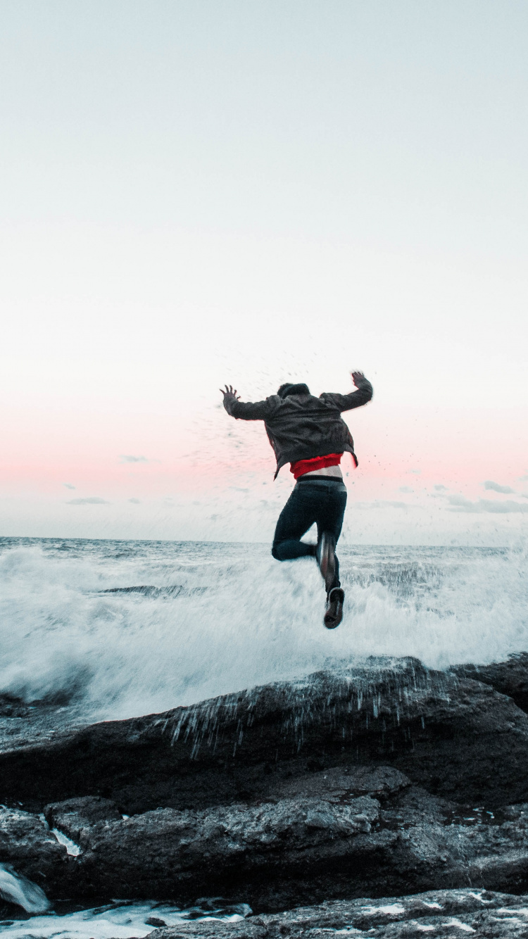 Man in Black Jacket and Pants Jumping on Rocky Shore During Daytime. Wallpaper in 750x1334 Resolution