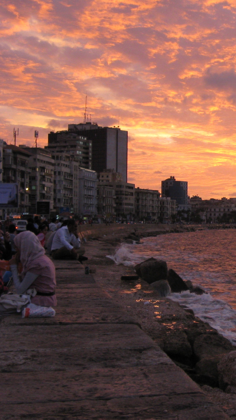 Hombre Sentado Sobre Una Roca Cerca Del Cuerpo de Agua Durante la Puesta de Sol. Wallpaper in 750x1334 Resolution