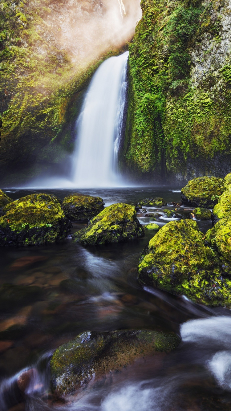 El Agua Cae en Medio de Rocas Cubiertas de Musgo Verde.. Wallpaper in 750x1334 Resolution