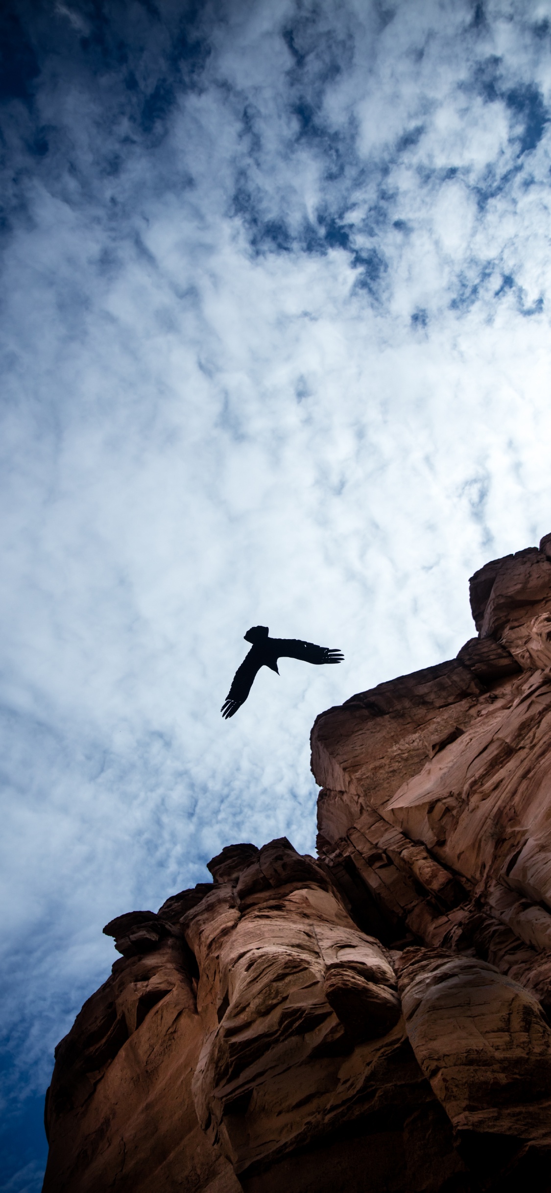 Man Jumping on Brown Rock Formation Sous Les Nuages Blancs et Ciel Bleu Pendant la Journée. Wallpaper in 1125x2436 Resolution