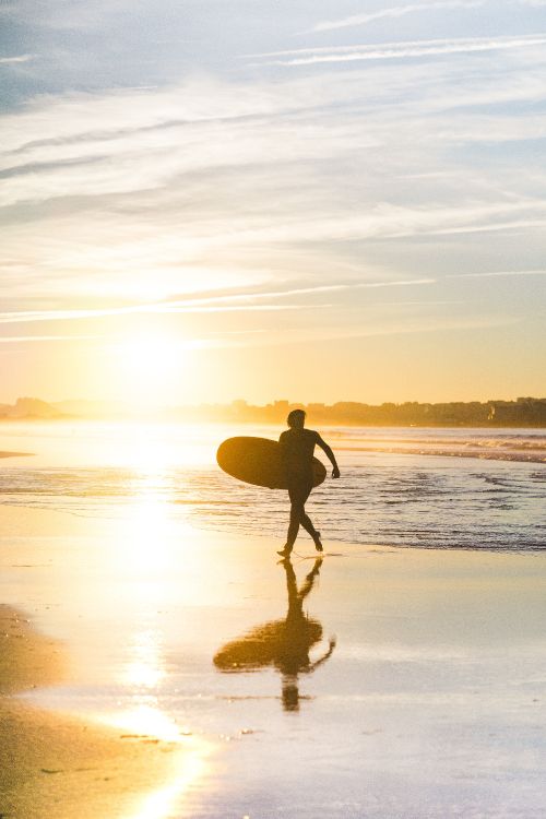 Hombre en Shorts Negros Sosteniendo la Tabla de Surf Caminando Por la Orilla Del Mar Durante la Puesta de Sol. Wallpaper in 4000x6000 Resolution