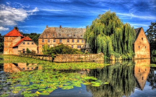 Image brown brick building near green trees and body of water under blue sky during daytime