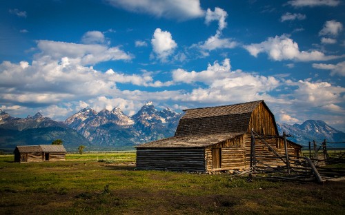 Image brown wooden barn on green grass field near snow covered mountains under blue and white cloudy