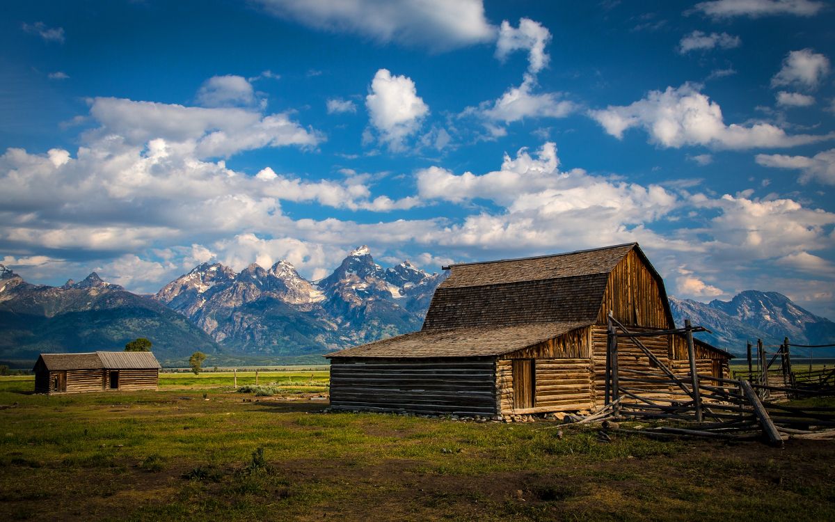 brown wooden barn on green grass field near snow covered mountains under blue and white cloudy