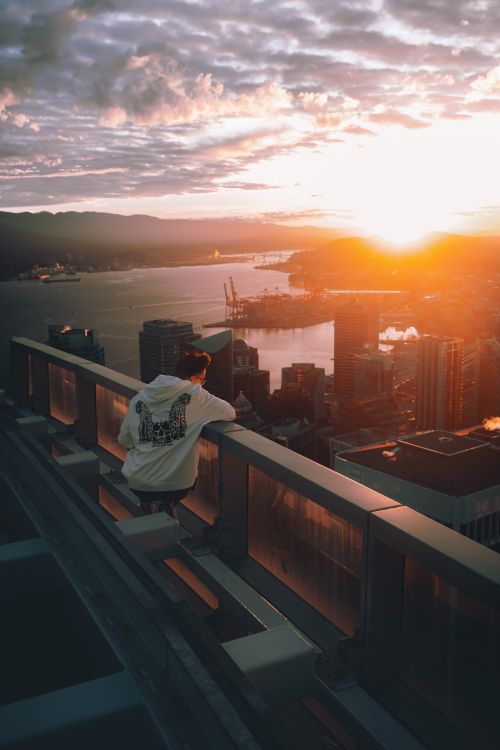 Homme en Sweat à Capuche Blanc Assis Sur Une Clôture en Bois Marron Regardant Les Toits de la Ville Pendant le Coucher du Soleil. Wallpaper in 3289x4933 Resolution
